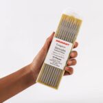 Hand holding of a 10-pack of Weldcraft 3.2mm (1/8 inch) gold pure tungsten electrodes for TIG welding, ground finish, on a gold background.